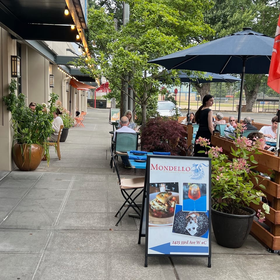 Mondello restaurant patio with outdoor dining, umbrellas, potted plants, and a sidewalk sign displaying food and drinks.