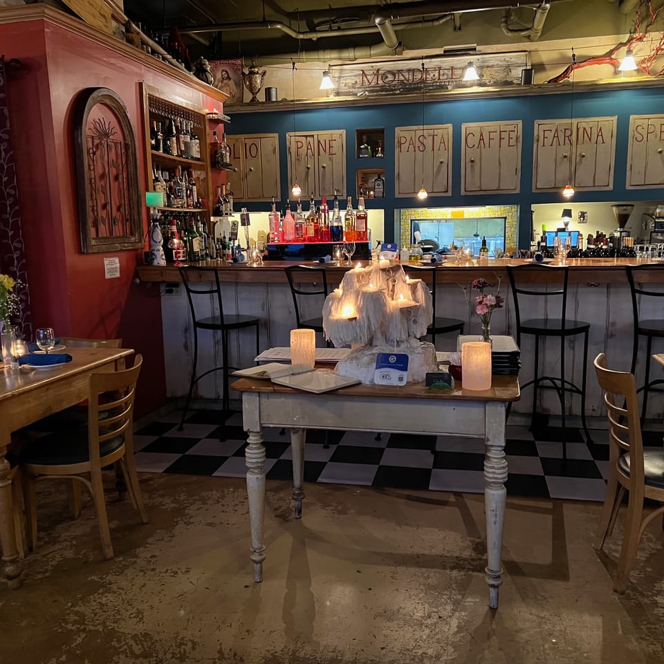 Warmly lit Italian restaurant interior with candlelit tables, a long bar, bottles, and wall signs labeled pane, pasta, caffè, farina, and spezie.