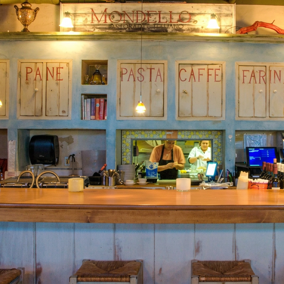 Rustic Italian restaurant bar with blue cabinetry, pendant lights, bottles, kitchen staff, and labeled cupboards reading olio, pane, pasta, and caffè.