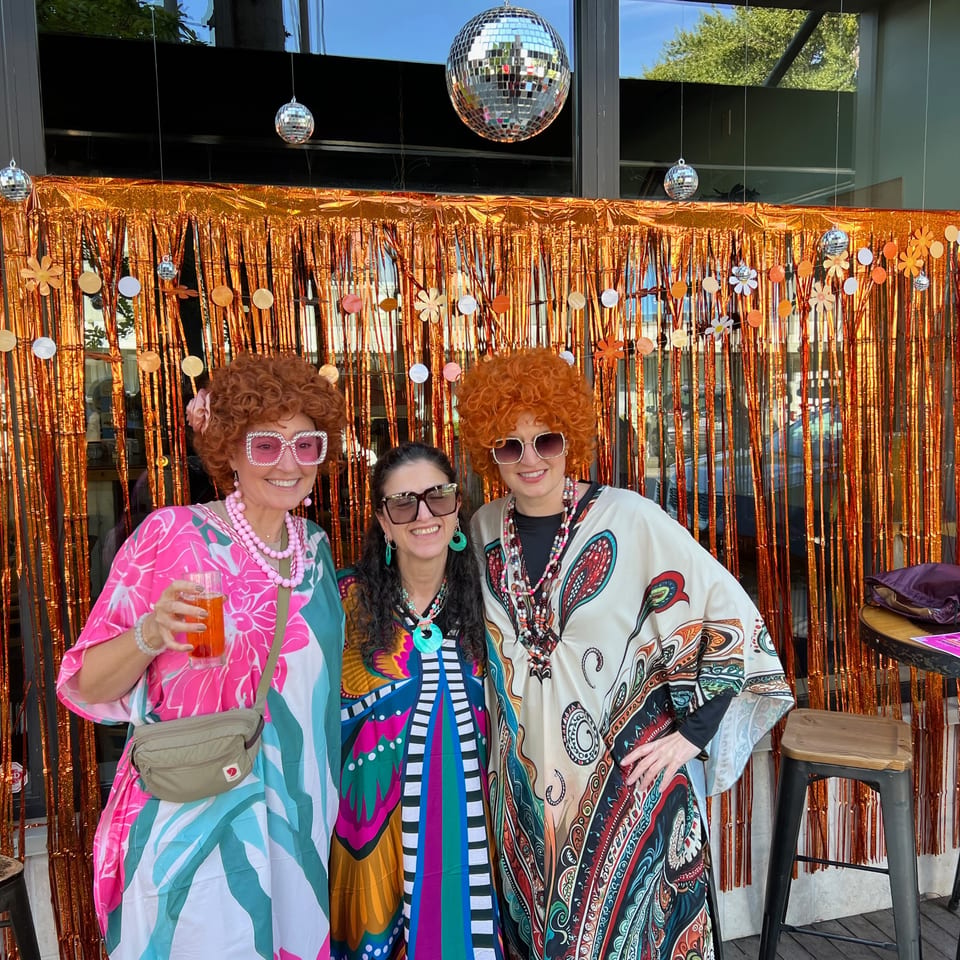 Three smiling women in colorful retro outfits pose before a shimmering orange tinsel backdrop with disco balls and floral decorations.