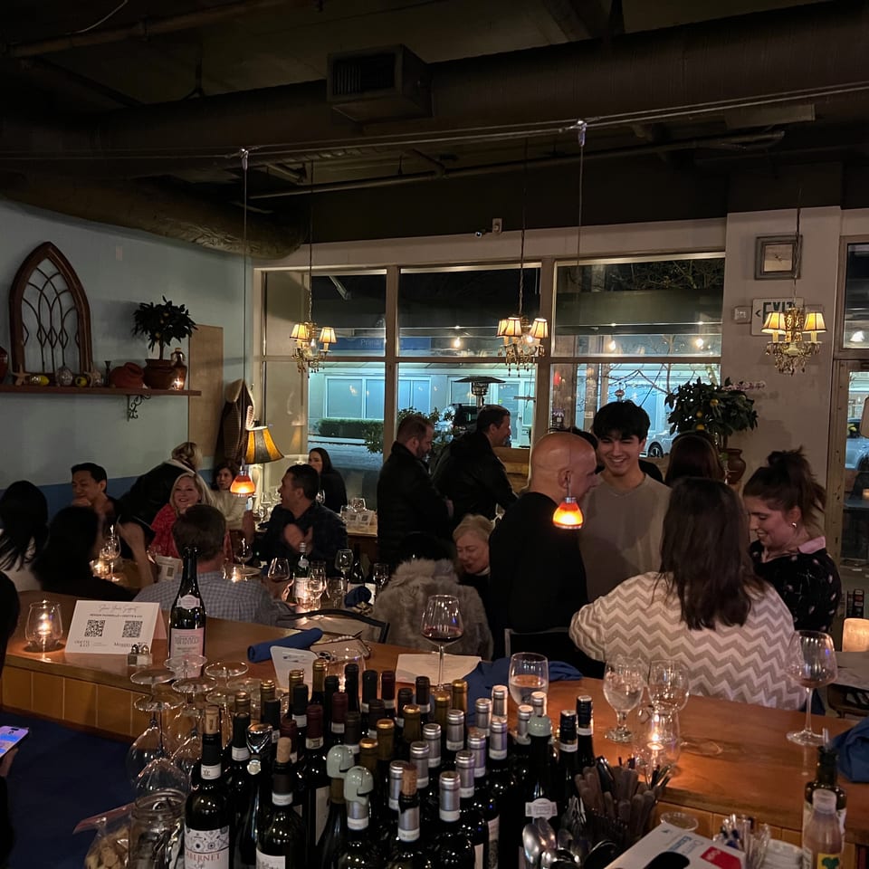 Bustling restaurant dining room filled with guests, candlelit tables, wine bottles, chandeliers, and an illuminated open sign.